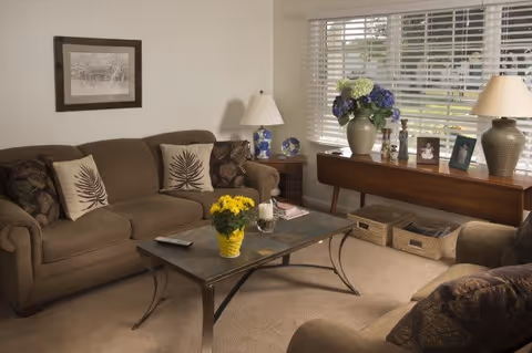 A cozy living room with a brown sofa adorned with decorative pillows, a glass-top coffee table with a yellow flower pot and a candle, a side table with a lamp and decorative plates, and a window with white blinds letting in natural light. There is also a wooden console table under the window with a large vase of flowers, framed photos, and two wicker baskets underneath.