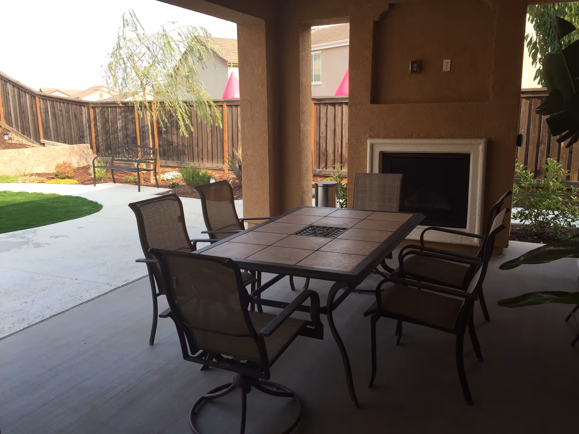 Covered outdoor patio area with a rectangular tiled table surrounded by six chairs. There is a built-in fireplace on the wall and a wooden fence enclosing a landscaped yard with a bench, plants, and a small tree.