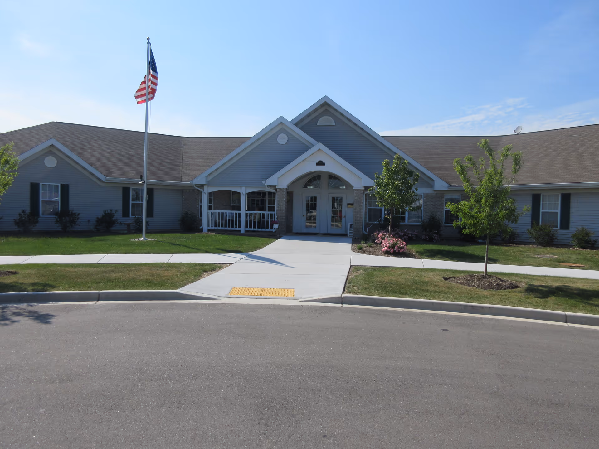 Front exterior view of a single-story building with gray siding and a peaked roof. There is a concrete walkway leading to double glass doors under a small covered porch. An American flag on a flagpole is positioned on the left side of the walkway. The lawn is neatly maintained with small trees and shrubs, and the sky is clear and blue.