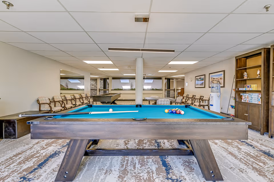 A recreational room with a pool table in the center, a shuffleboard table in the background, several chairs lined up against the walls, a water cooler, and a bookshelf with board games and decorative items. The room has a patterned carpet and a drop ceiling with fluorescent lighting.