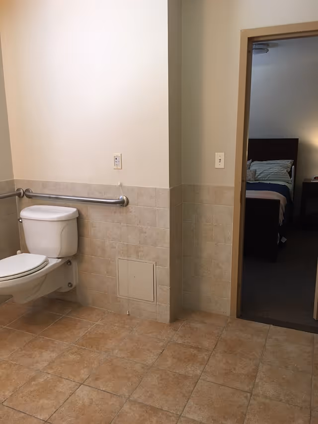 Accessible bathroom with a wall-mounted toilet, horizontal grab bar, tiled walls and floor, and a doorway leading to a bedroom.
