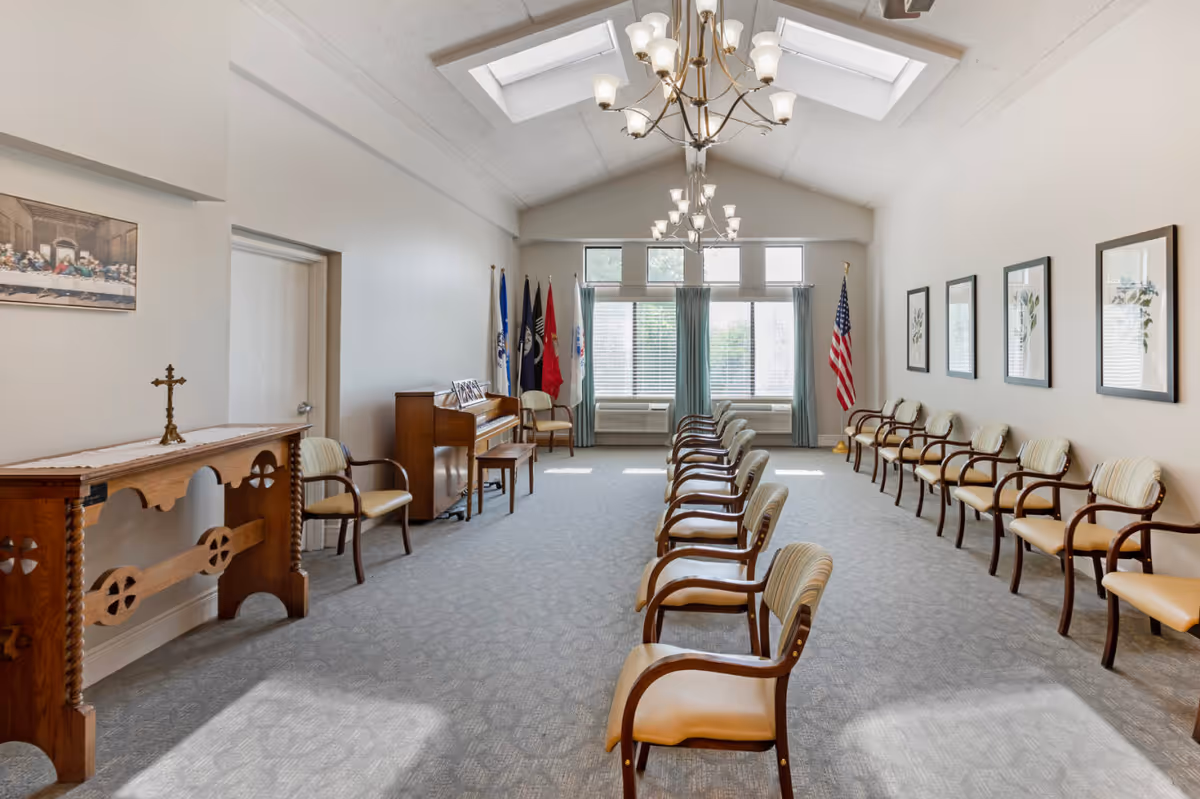 Bright interior meeting/chapel room with two rows of chairs facing each other, a piano, flags, and a cross on a wooden table.