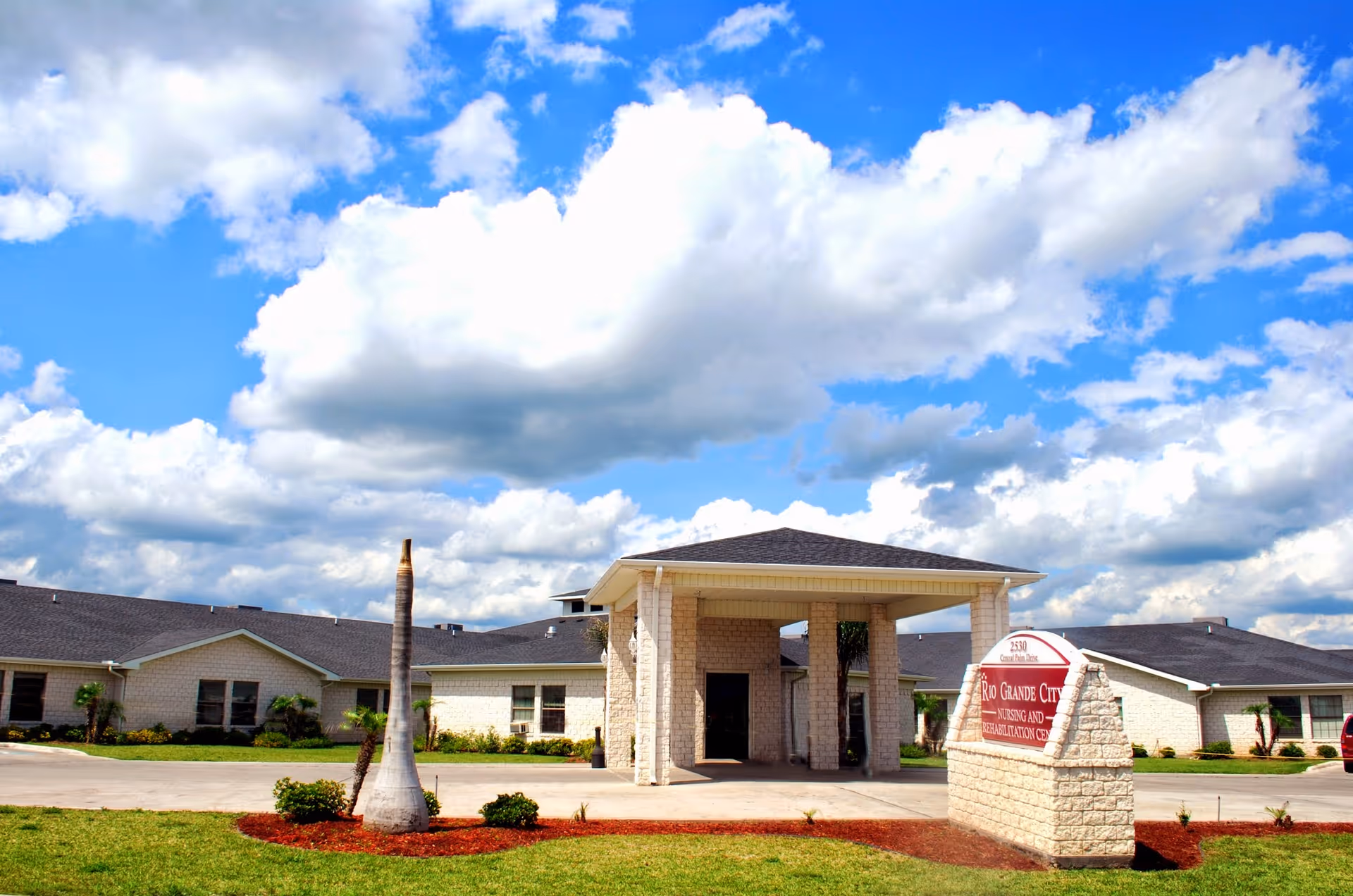Front entrance of a single-story nursing and rehabilitation center with a porte-cochère, sign, small palm trees, and a blue sky with clouds.