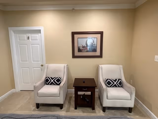 Two beige armchairs with black and white patterned pillows are placed on either side of a small dark wooden table with a drawer and a remote control on top. Behind the chairs is a beige wall with a framed picture hanging above the table. To the left is a white door with the number 2 on it.