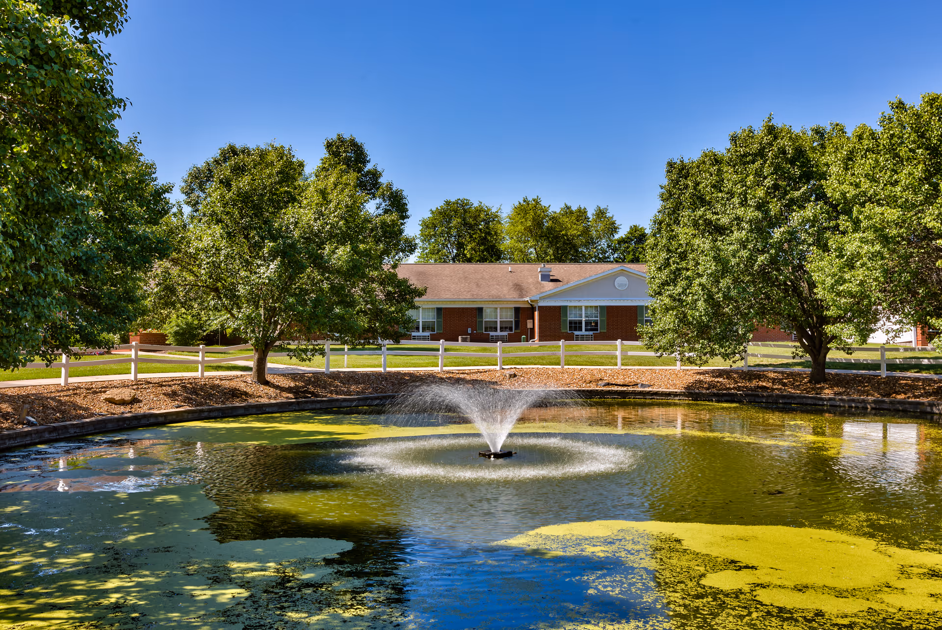 A pond with a central fountain and algae in front of a single-story brick building flanked by trees and a white fence.