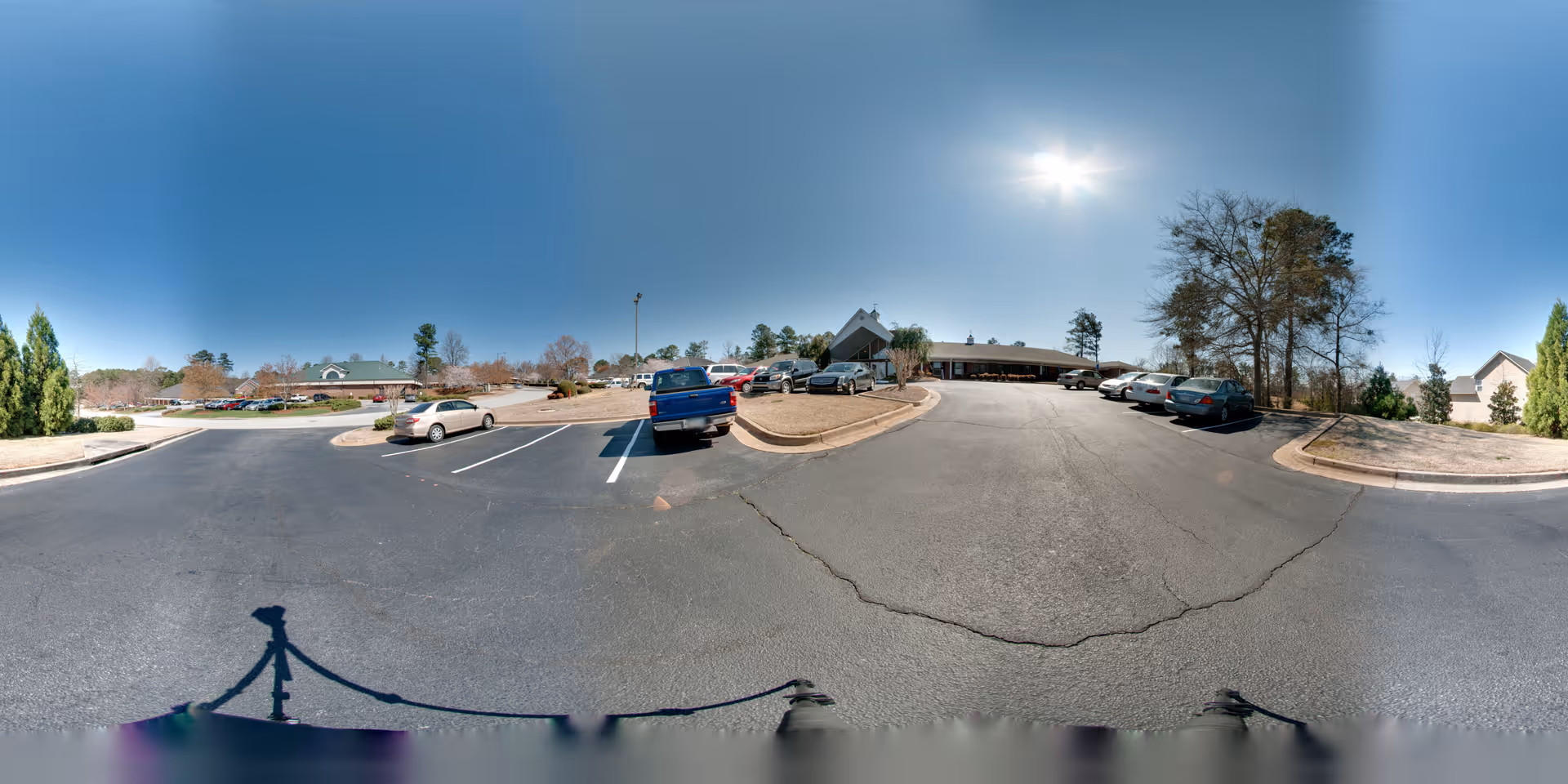 Wide panoramic view of a senior living facility parking lot on a sunny day with several parked cars, trees, and the main building entrance visible in the background under a clear blue sky.