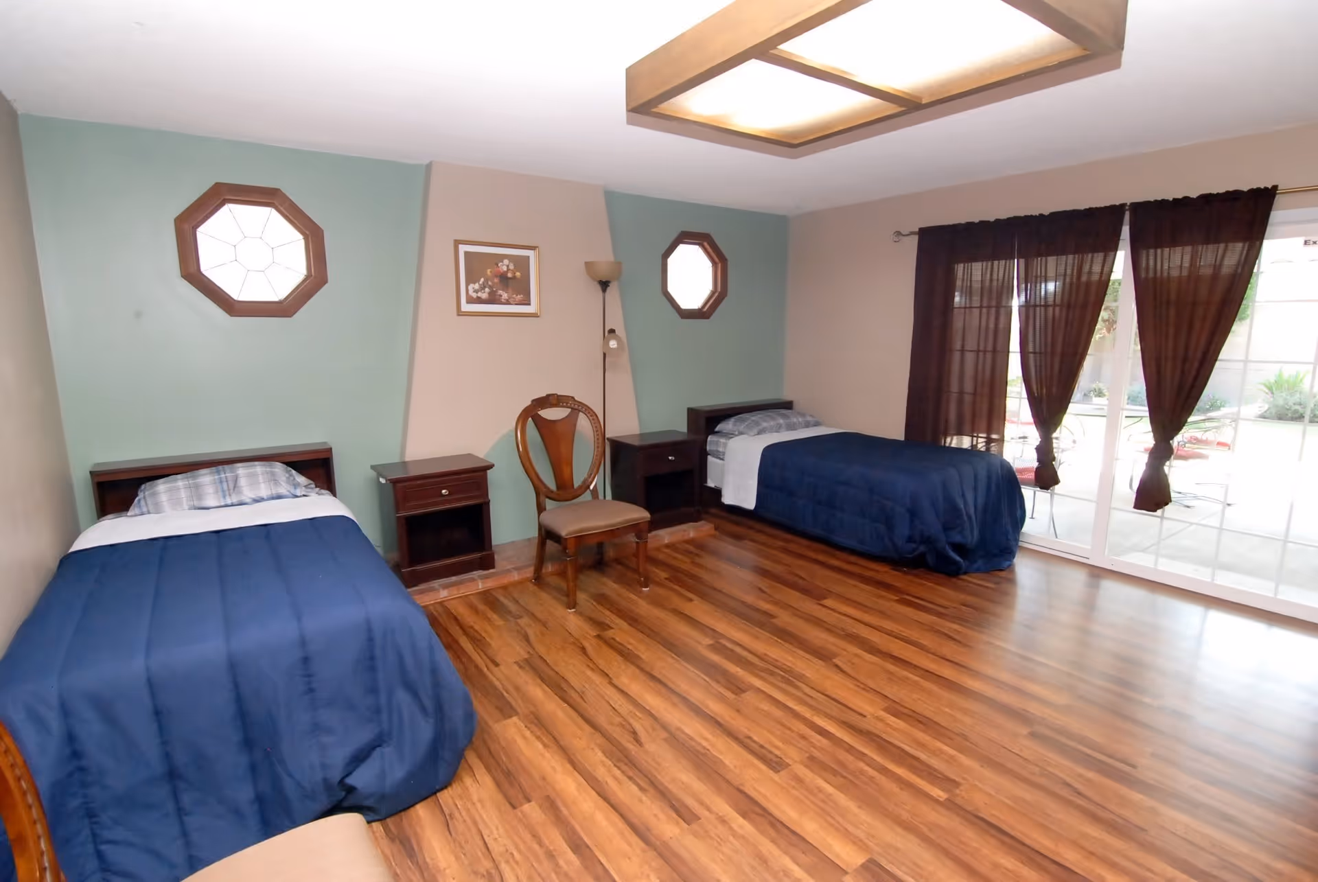 Spacious bedroom with two twin beds, nightstands, octagonal windows, wood flooring, and sliding glass doors with dark curtains.