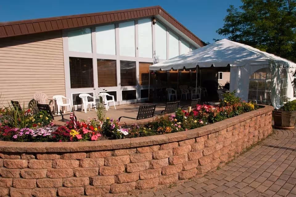Raised brick flowerbed and paved patio with chairs and a white canopy tent outside a light-colored building.