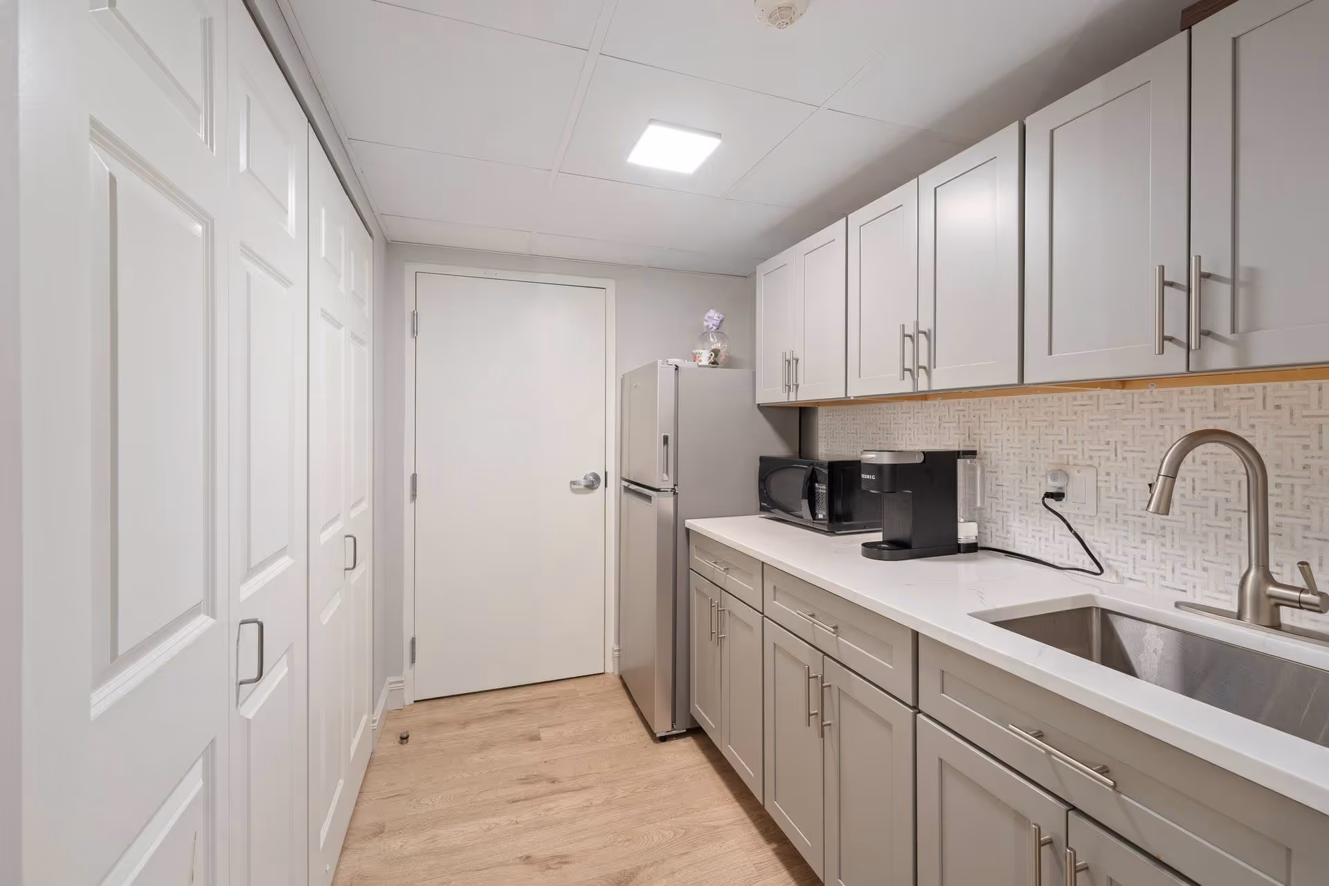 A modern kitchen with light gray cabinets, a stainless steel refrigerator, a microwave, a coffee maker, a stainless steel sink with a faucet, and a white countertop. The floor is light wood, and there is a white door at the end of the room. The backsplash has a textured, patterned design.