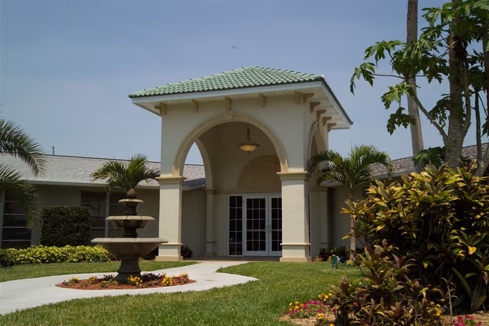 Entrance of a light-colored building with an arched portico, green-tiled roof, fountain and landscaped lawn.