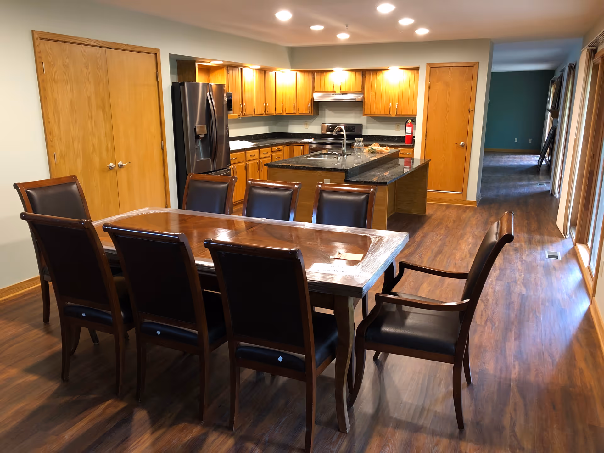 A dining area with a long wooden table surrounded by eight chairs with dark cushions. Behind the dining area is a kitchen with wooden cabinets, a black countertop island with a sink, a stainless steel refrigerator, and a stove. The floor is wooden, and there are two wooden doors on the left and right sides of the kitchen. The room is well-lit with recessed ceiling lights.