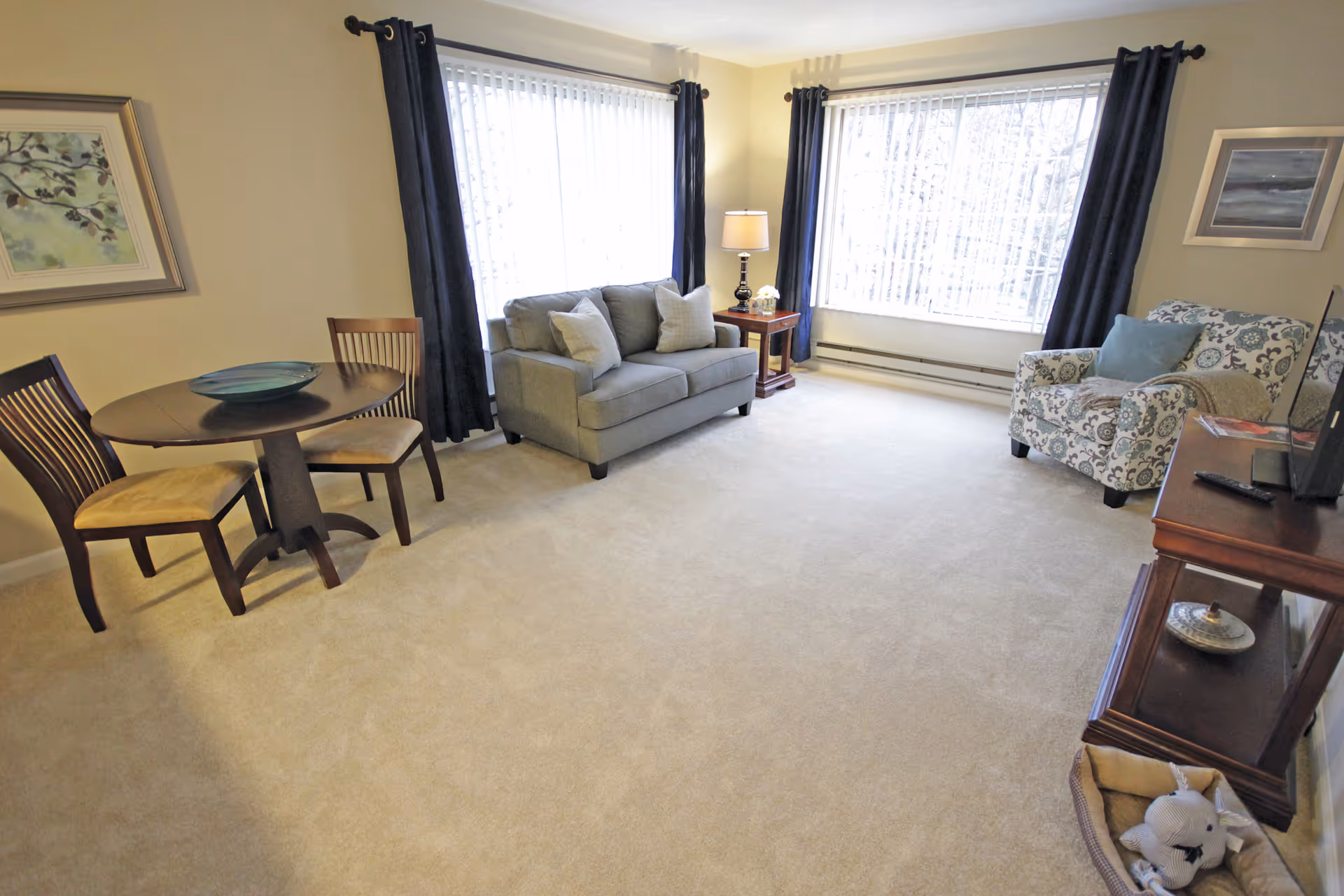 Bright furnished living room with a sofa, patterned armchair, round dining table, TV stand and large windows with curtains.