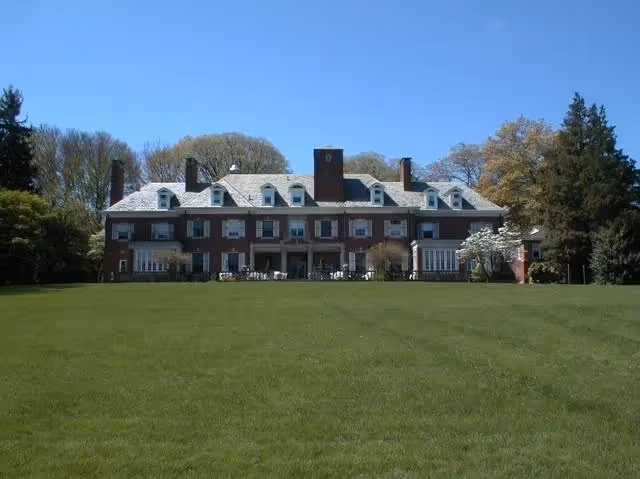 A large, multi-story brick building with multiple chimneys and dormer windows, surrounded by trees and a wide green lawn under a clear blue sky.