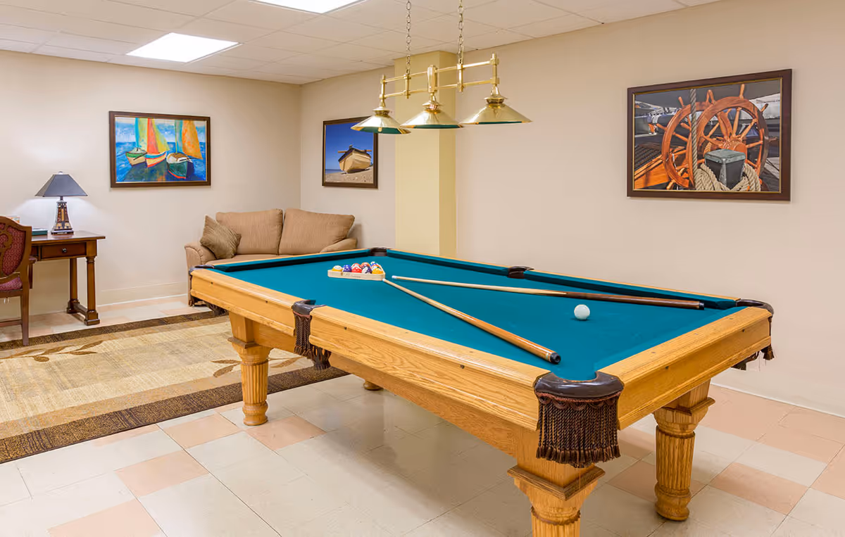 A well-lit recreation room with a wooden pool table and cues, a sofa and desk, and framed nautical artwork on the walls.