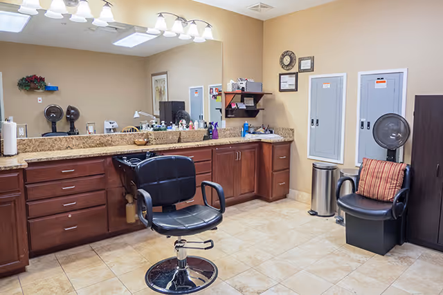 Salon-style room with a black hydraulic styling chair, sink, large mirror, and wooden cabinets against a tiled floor.