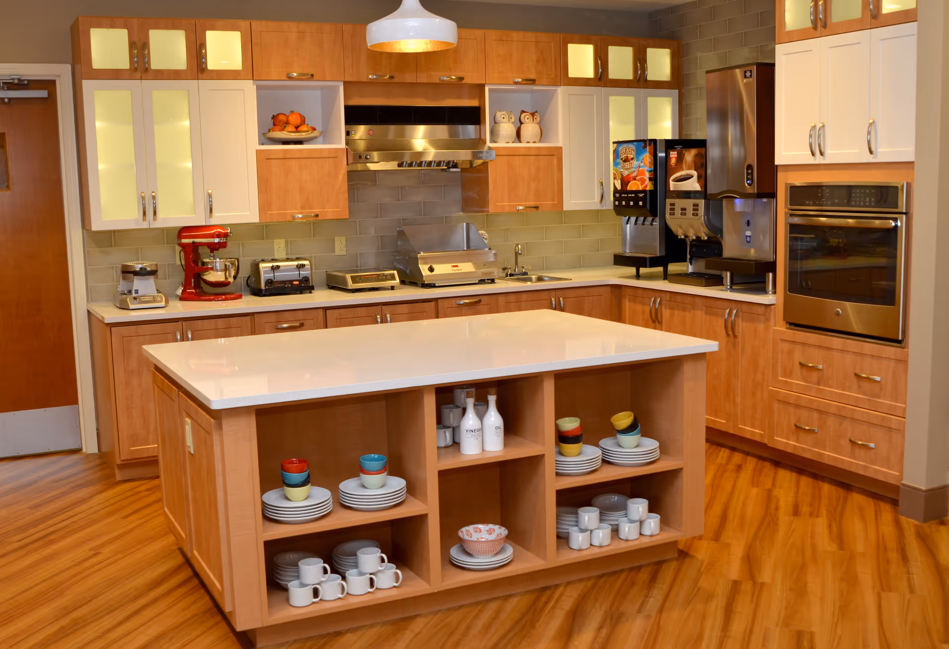 A modern kitchen with wooden cabinets and a large central island with open shelves holding plates, cups, and bowls. The kitchen features stainless steel appliances including an oven, a toaster, a coffee machine, and a mixer. The floor is wooden, and there is a white pendant light hanging above the island.