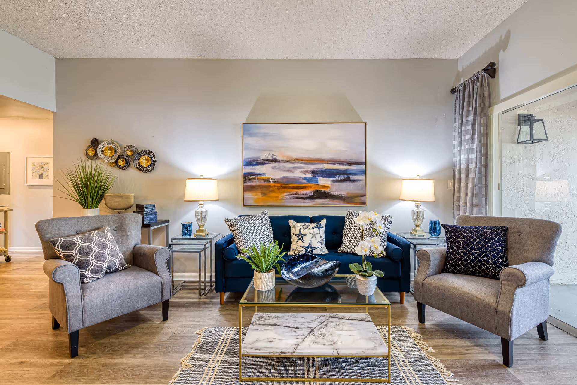 Bright furnished living room with a blue sofa flanked by two gray armchairs, a glass coffee table, lamps, and abstract wall art.