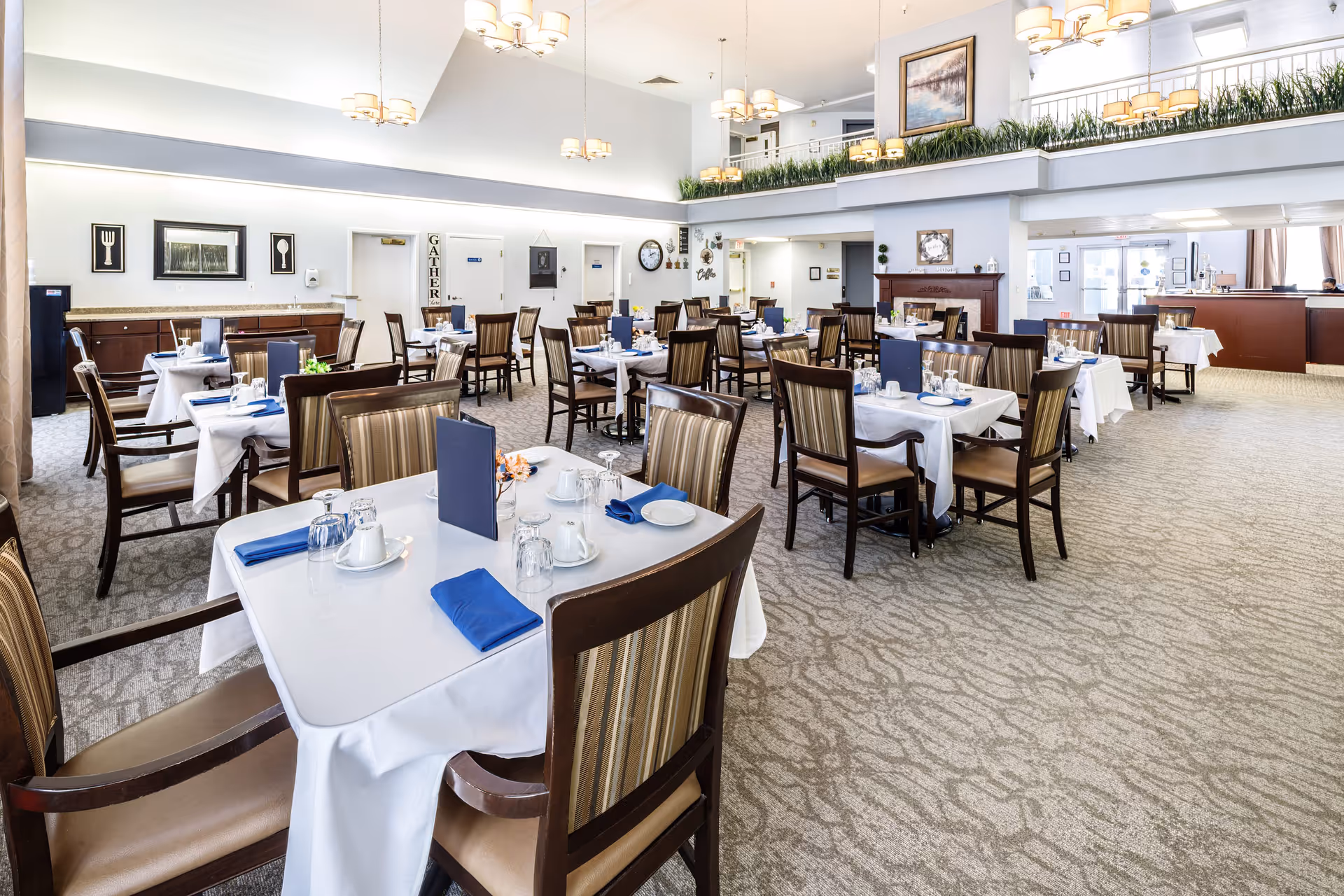 A spacious and well-lit dining room in Parkway Assisted Living and Memory Care, featuring multiple tables covered with white tablecloths, each set with plates, cups, glasses, and blue napkins. The room has high ceilings with modern chandeliers, striped cushioned chairs, framed artwork on the walls, and a carpeted floor. There is a counter area in the background and a second level with greenery along the railing.