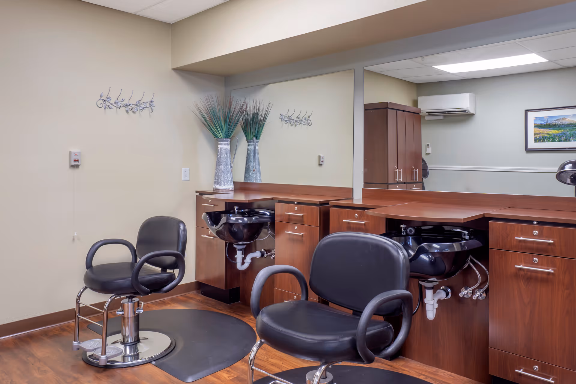 Interior of a hair salon area with two black salon chairs in front of wooden counters with built-in black sinks. A large mirror spans the wall behind the counters. Decorative vases with tall grass and a wall-mounted artwork are visible. The floor is wooden, and the ceiling has recessed lighting.