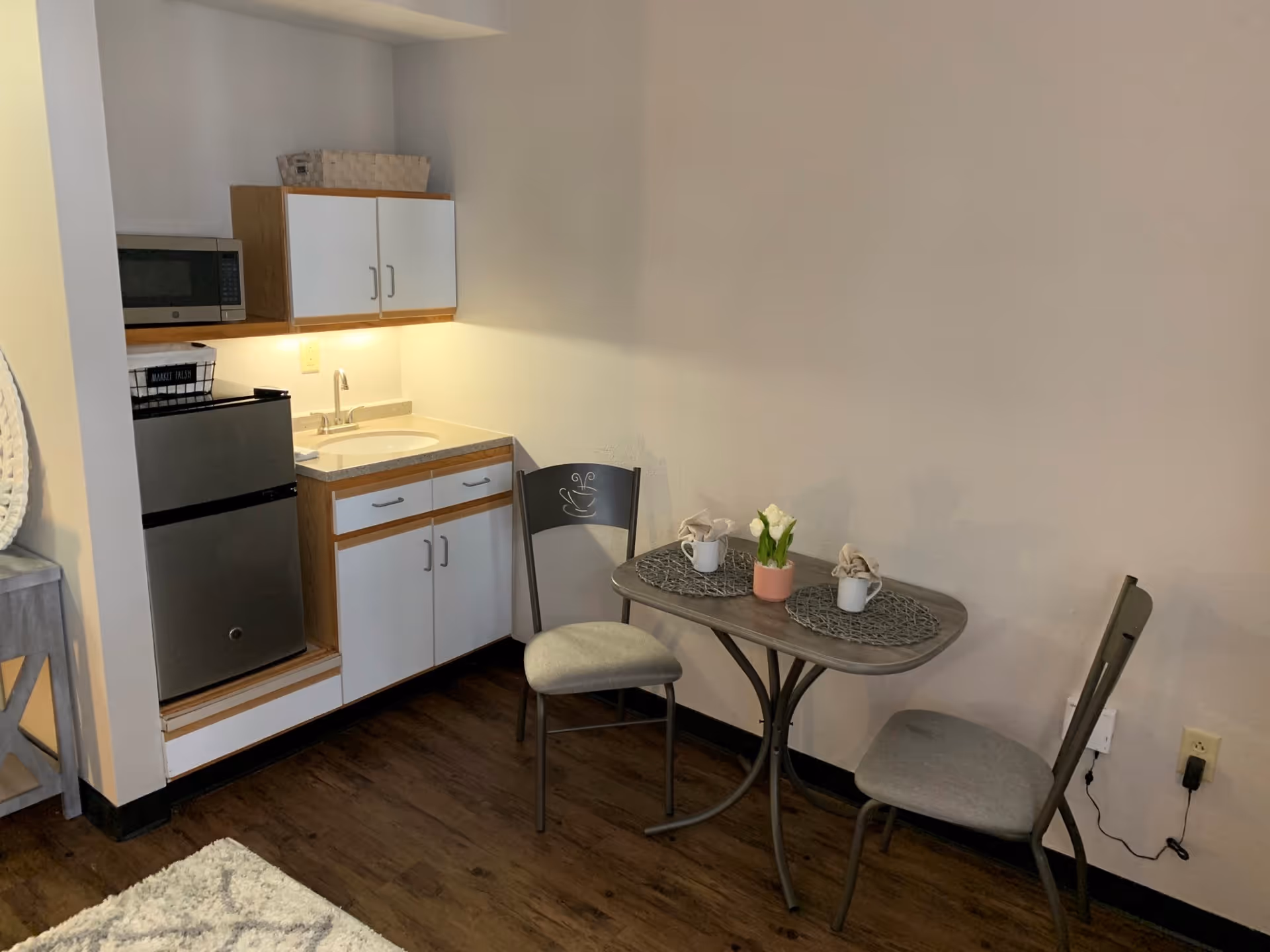 Small kitchenette area with a mini refrigerator, microwave, sink, and cabinets. Next to the kitchenette is a small dining table with two chairs, each with a placemat, a mug, and a napkin. A small potted plant with white flowers is on the table. The floor is wooden and the walls are light-colored.