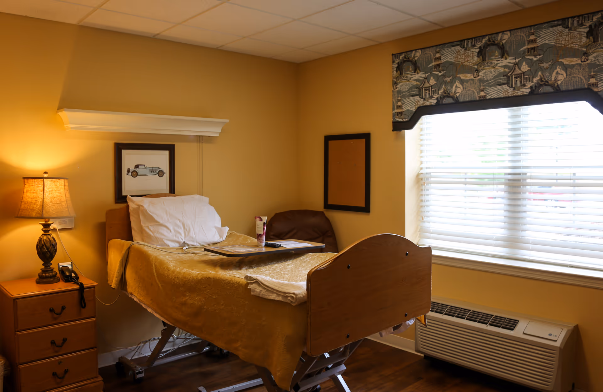A cozy patient room in a healthcare facility with a single adjustable bed covered with a gold blanket and white pillows. Next to the bed is a wooden nightstand with a lamp and a telephone. A framed picture of a vintage car hangs on the wall above the bed. A window with blinds and a decorative valance lets in natural light. There is also a brown chair and a heating/cooling unit beneath the window.