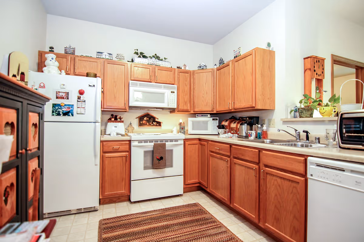 A cozy kitchen with wooden cabinets, a white refrigerator, a white stove with a microwave above it, a dishwasher, and a sink. The countertops have various kitchen appliances and decorative items. There is a small rug on the tiled floor and a wooden clock on the wall near a pass-through window.