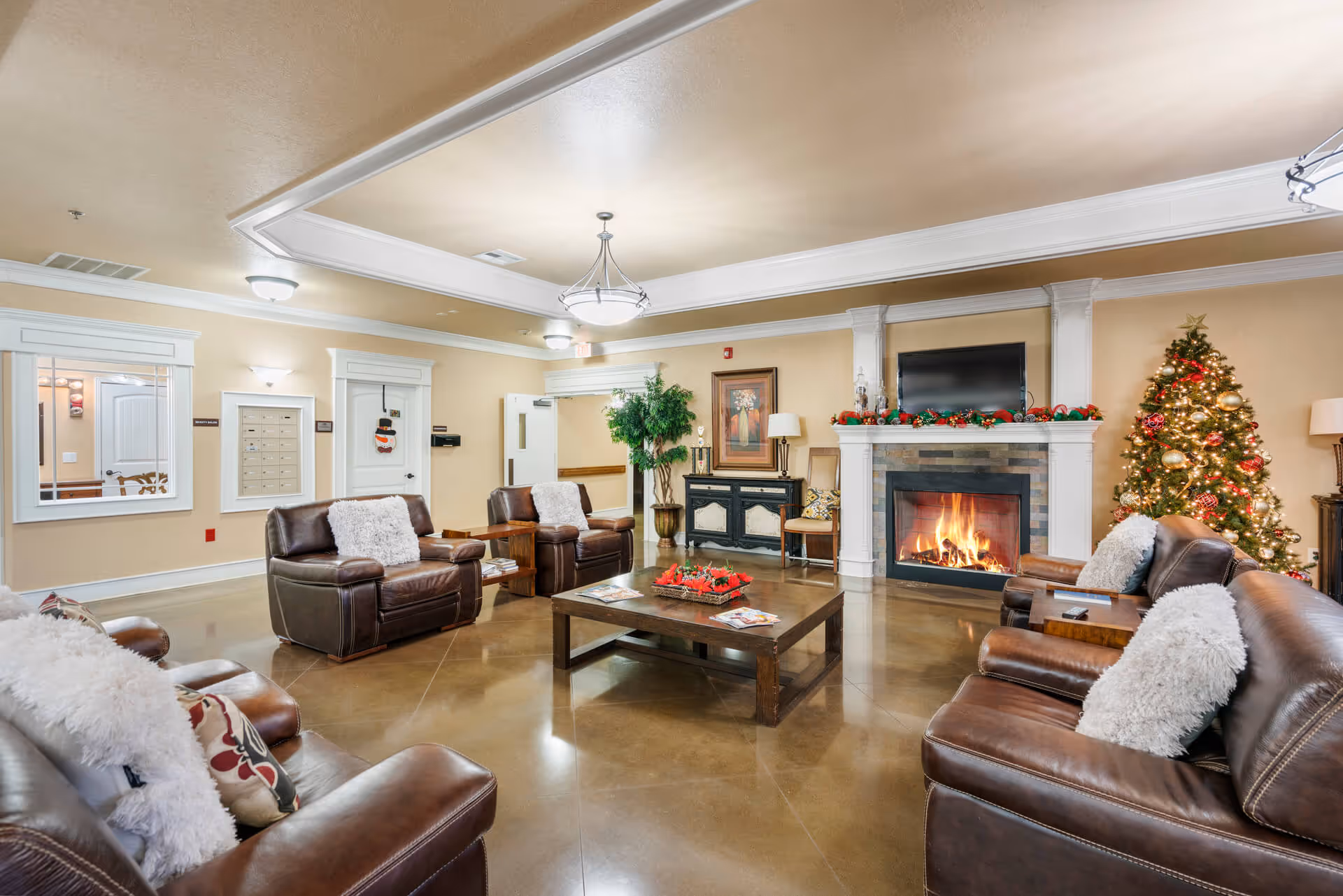A cozy senior living facility common area with leather sofas and armchairs arranged around a wooden coffee table. The room features a lit fireplace decorated with Christmas garlands, a Christmas tree with ornaments and lights, a flat-screen TV mounted above the fireplace, and warm beige walls with white trim. There is a large plant and framed artwork on the wall, with soft lighting from ceiling fixtures.