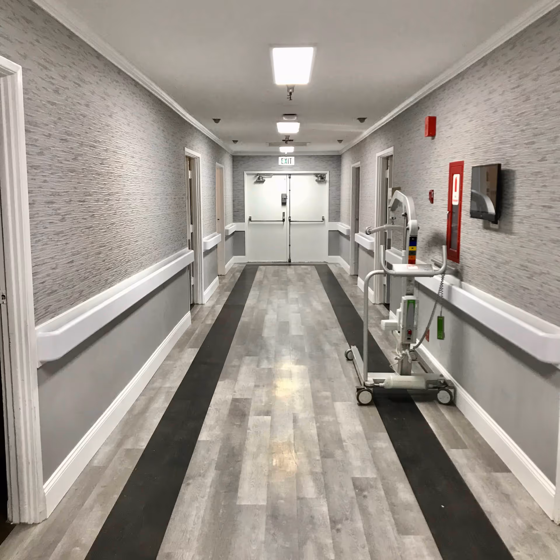 A clean, well-lit hallway in a senior living facility with light gray walls, white trim, and a gray and black patterned floor. There are several doorways along the hallway and a white medical lift device on the right side near a fire alarm and a mounted screen. Double exit doors are visible at the end of the hallway.