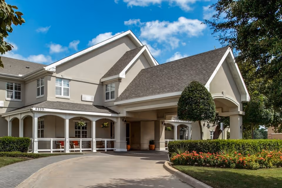 Exterior view of a senior living facility building with beige walls and a gray shingled roof under a blue sky with scattered clouds. The entrance features a covered driveway with white pillars, surrounded by neatly trimmed bushes and colorful flowers.