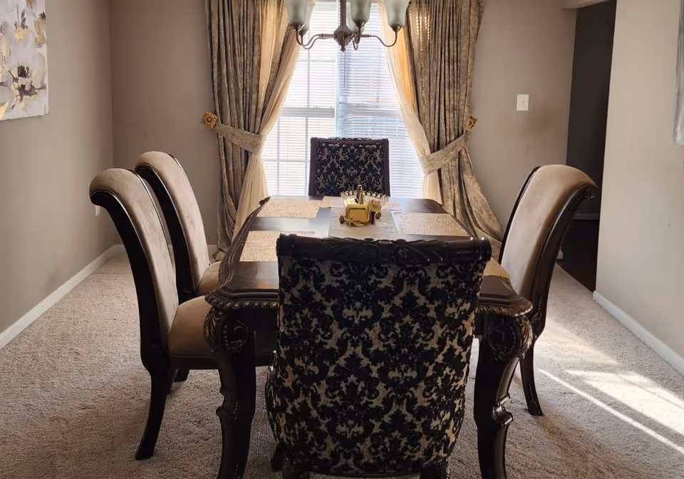 A dining room with a dark wooden table and six chairs, two of which have a black and beige patterned upholstery. The room has beige carpet, beige walls, and a window with beige curtains tied back, allowing sunlight to enter. A chandelier hangs above the table.