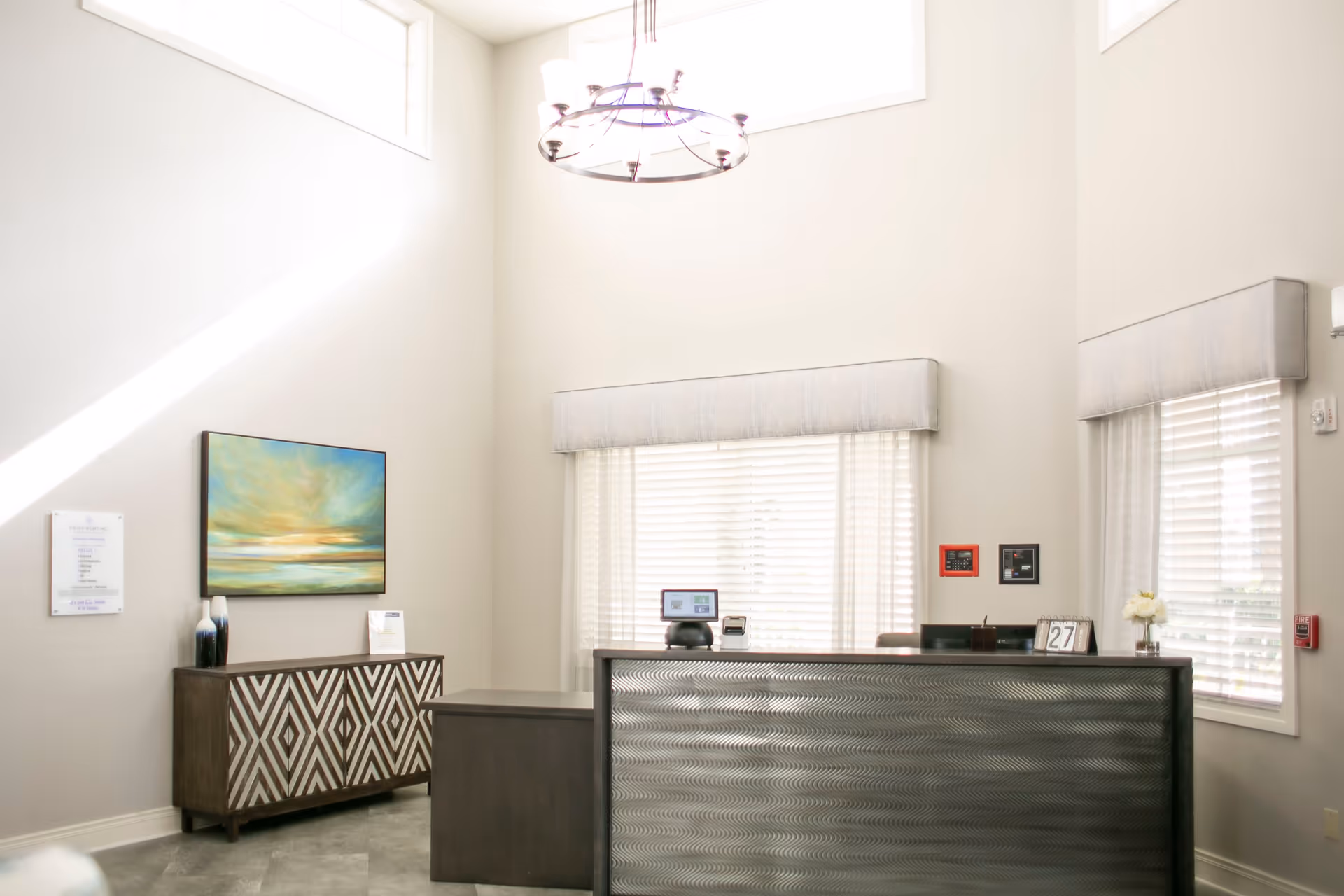 Bright reception area with a modern desk featuring a textured front panel, two large windows with blinds and valances, a decorative sideboard with geometric patterns, a colorful landscape painting on the wall, and a circular chandelier hanging from a high ceiling.
