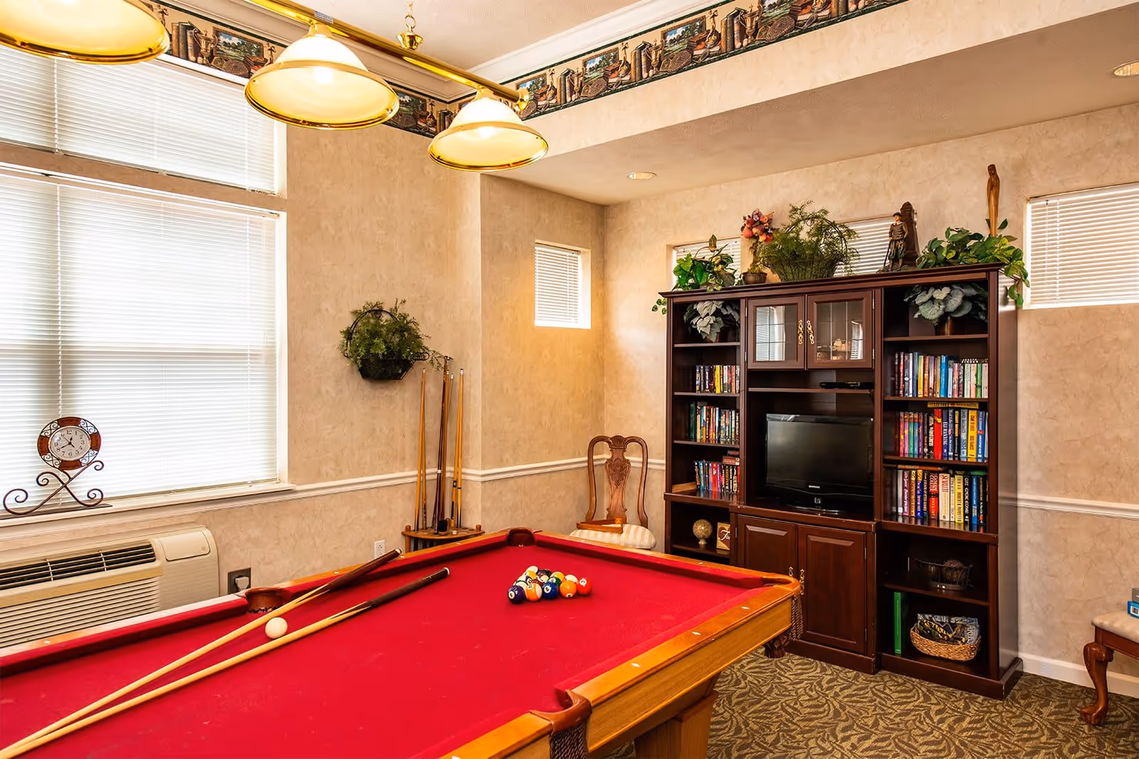 A cozy game room featuring a red felt pool table with pool cues resting on it. The room has beige walls with a decorative border near the ceiling, two windows with blinds, and a dark wooden entertainment center filled with books, plants, and a small television. There is also a wooden chair with a cushioned seat and a wall-mounted plant.