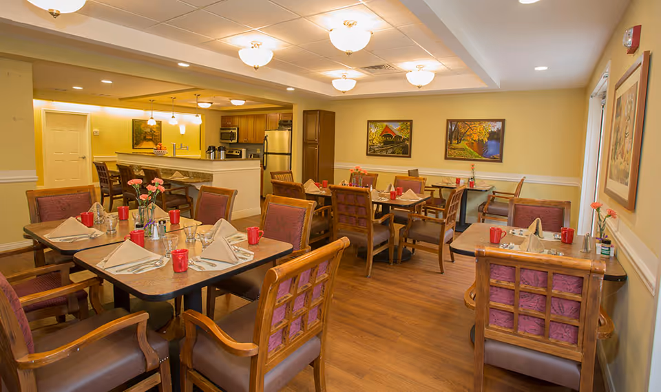 A cozy dining room in a senior living facility with several tables set with napkins, utensils, red mugs, and small flower vases. The room has wooden chairs with cushioned seats and backs, warm yellow walls, wood flooring, and framed landscape paintings on the walls. In the background, there is a kitchen area with stainless steel appliances and a counter with bar stools.