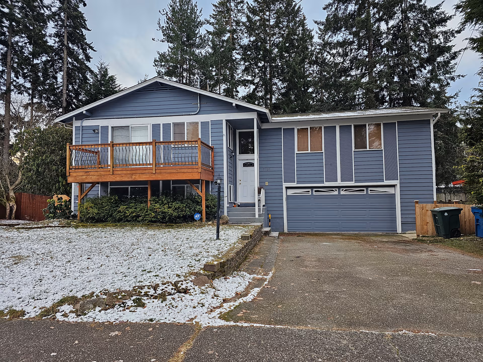 Blue split-level house with a wooden balcony, attached two-car garage, driveway and a light dusting of snow on the lawn.
