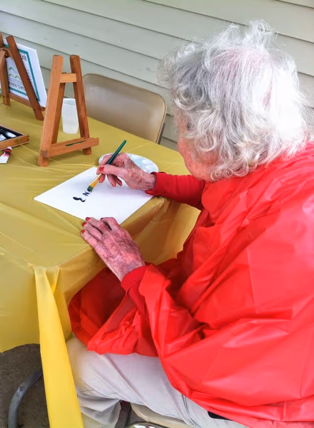 An elderly person with white hair wearing a red jacket is seated at a yellow table, painting on a white sheet of paper using a paintbrush. There is a small wooden easel and a chair in the background.