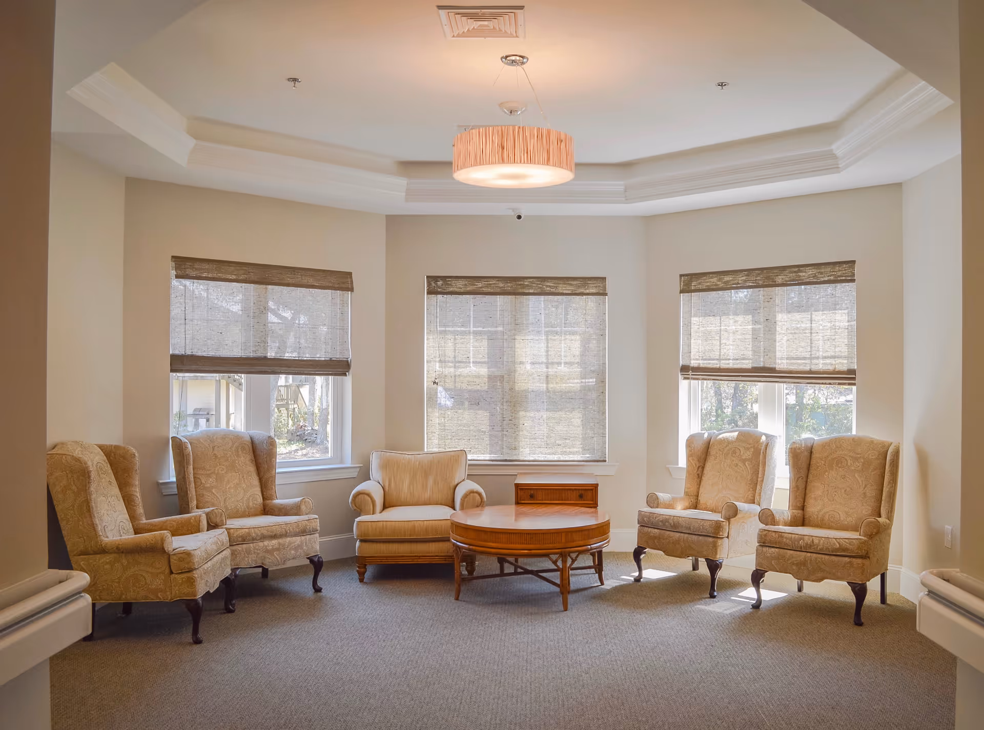 Sunlit seating area with five upholstered armchairs arranged around a round wooden coffee table in front of three large windows.
