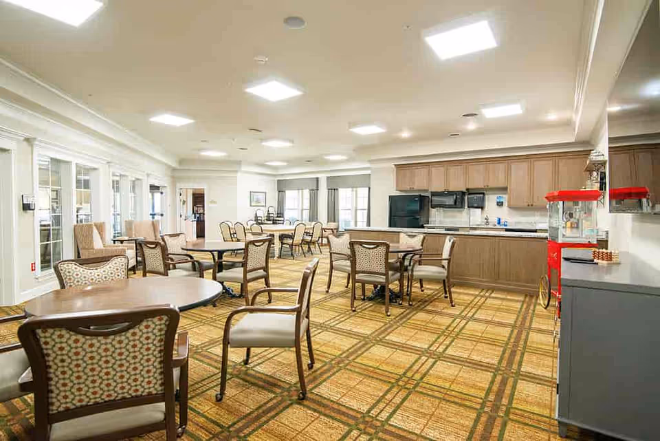 Well-lit communal dining area with multiple round tables and chairs and a kitchenette along the back wall.