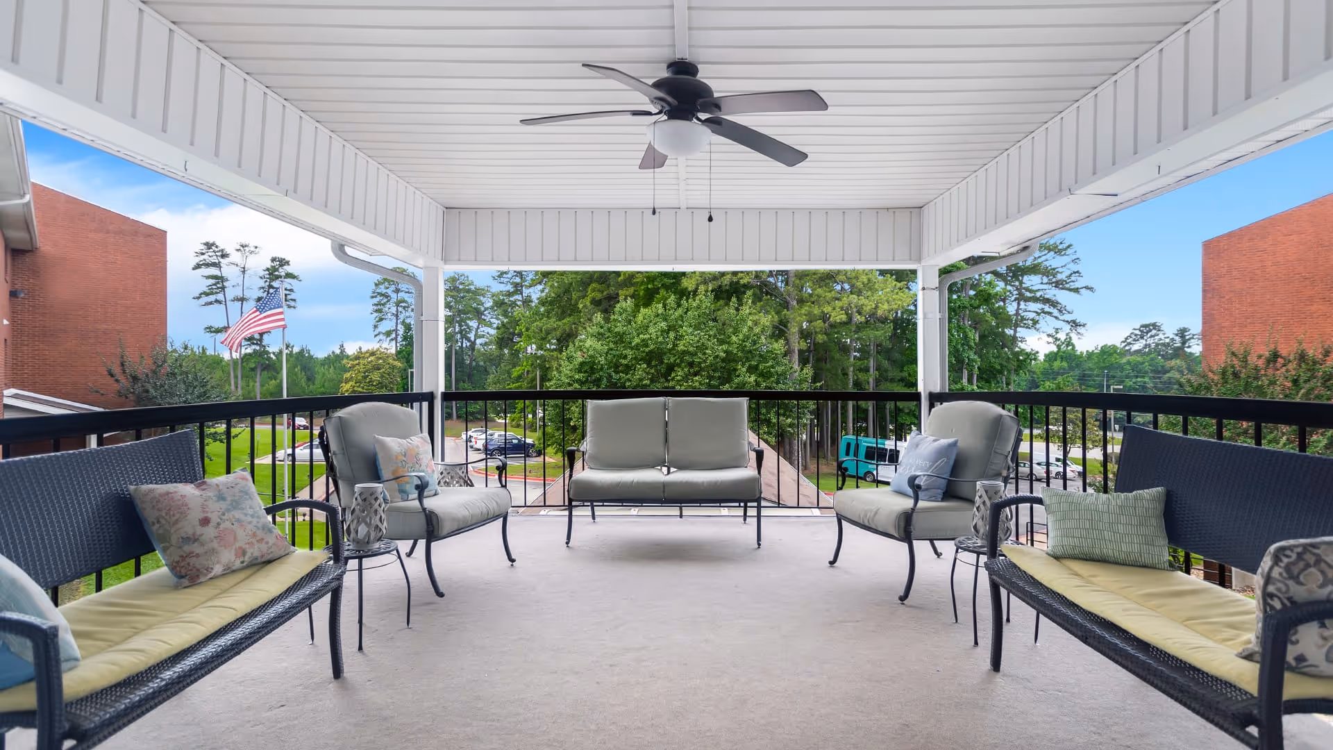 Covered outdoor patio area with cushioned seating including two benches and two armchairs arranged around small side tables. A ceiling fan is mounted on the white paneled ceiling. The patio overlooks a parking area with cars, green trees, and two red brick buildings on either side. An American flag is visible on the left side.