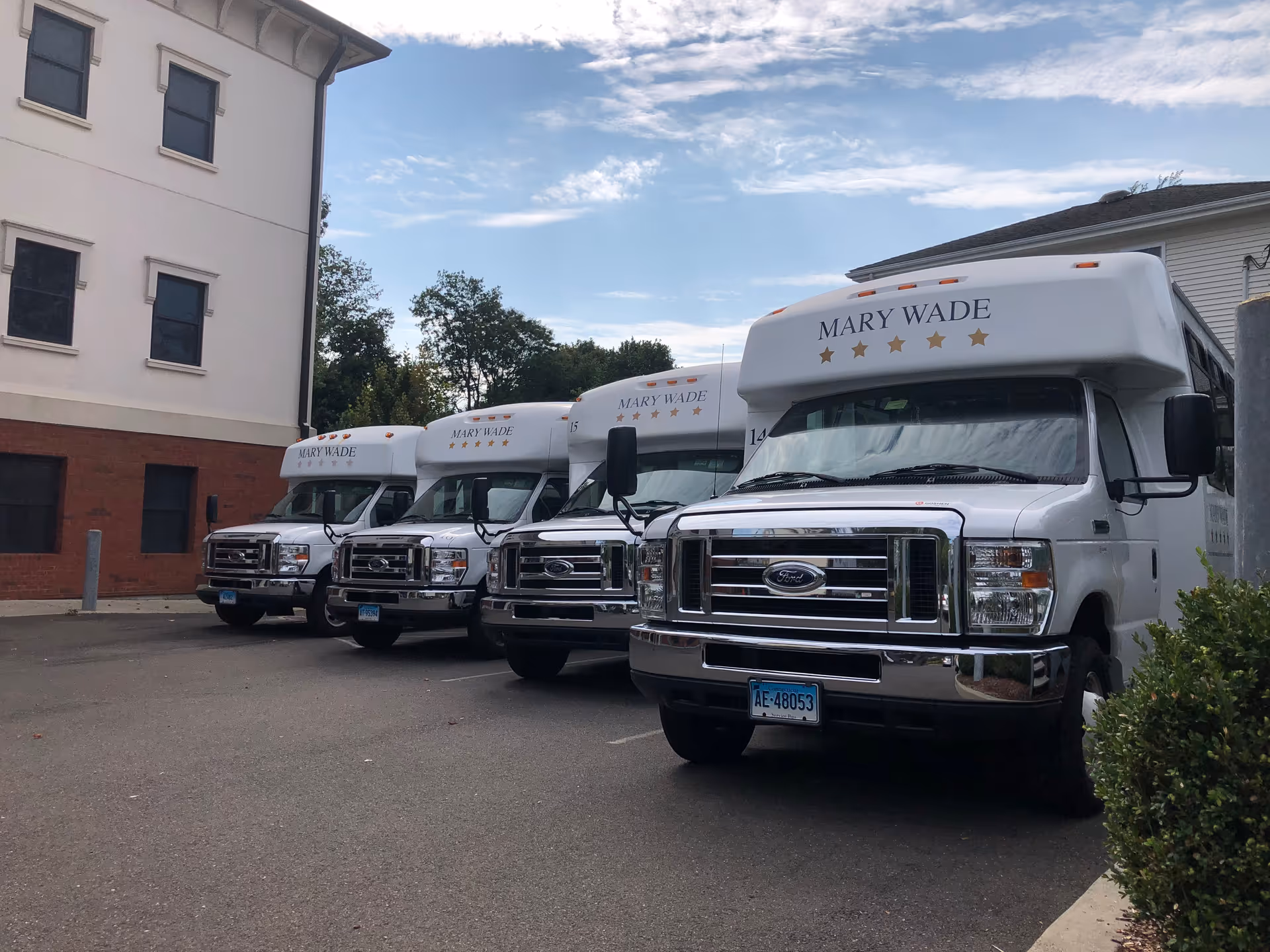 Four white shuttle buses parked side by side outside a building under a partly cloudy sky. Each bus has the name 'MARY WADE' and five gold stars displayed on the front above the windshield.
