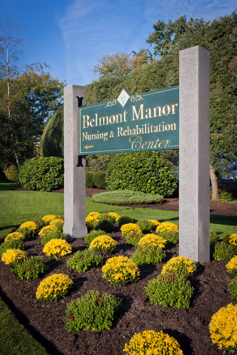 A green and gold sign for Belmont Manor Nursing & Rehabilitation Center mounted on two stone pillars, surrounded by neatly landscaped flower beds with yellow and green plants, with trees and a blue sky in the background.