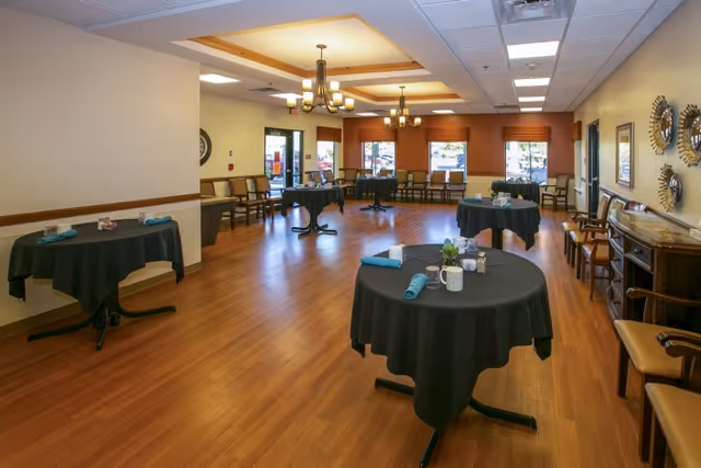 A spacious dining room with several round tables covered with black tablecloths, each set with teal napkins and small centerpieces. The room has wooden flooring, beige walls, multiple chairs lined up along the walls, and large windows with orange valances letting in natural light. Ceiling lights with chandeliers illuminate the room.