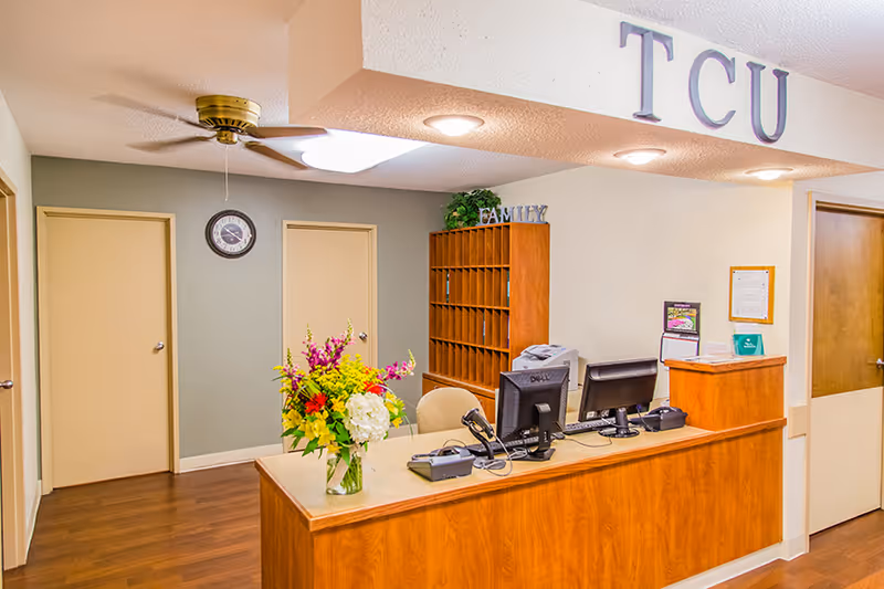 Reception desk area in a healthcare facility with two computer monitors, a telephone, and a vase of colorful flowers on the counter. Behind the desk is a wooden mail organizer with the word FAMILY on top. The walls are light-colored with two closed doors and a clock mounted on the wall. The ceiling has a fan and recessed lighting, and large letters spelling TCU are mounted above the reception area.