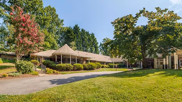 Exterior view of a single-story building with a covered entrance, surrounded by green grass, trees, and shrubs under a clear blue sky.
