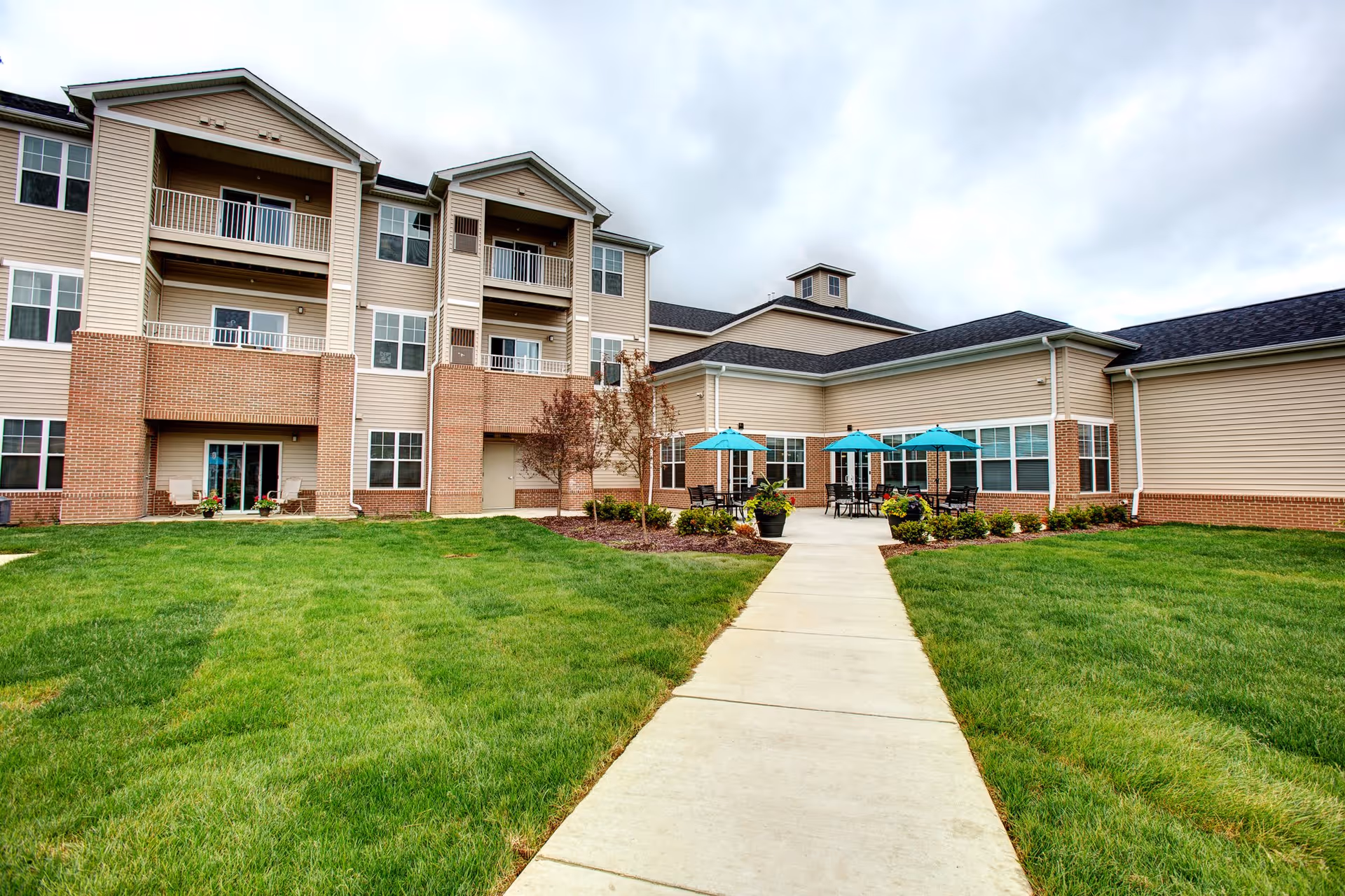 Exterior view of a multi-story senior living facility building with beige siding and brick accents. There is a concrete pathway leading to a patio area with tables and chairs under blue umbrellas. The surrounding area has well-maintained green grass and some small trees and shrubs.