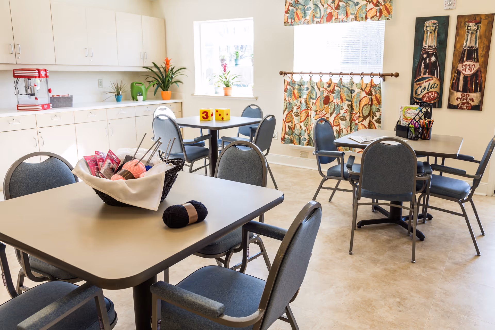 A bright room with several tables and chairs arranged for group activities. One table has a basket with yarn and knitting needles, and another table has large foam dice. The room has white cabinets along one wall, a popcorn machine, potted plants on the counter and windowsill, colorful patterned curtains, and wall art featuring vintage soda bottles.
