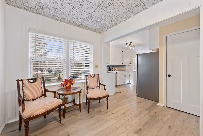 Bright sitting area with two upholstered wooden chairs and a small round table by windows, opening into a white kitchen.