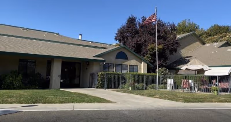 Single-story beige retirement community building with an American flagpole and a fenced patio under a clear blue sky.