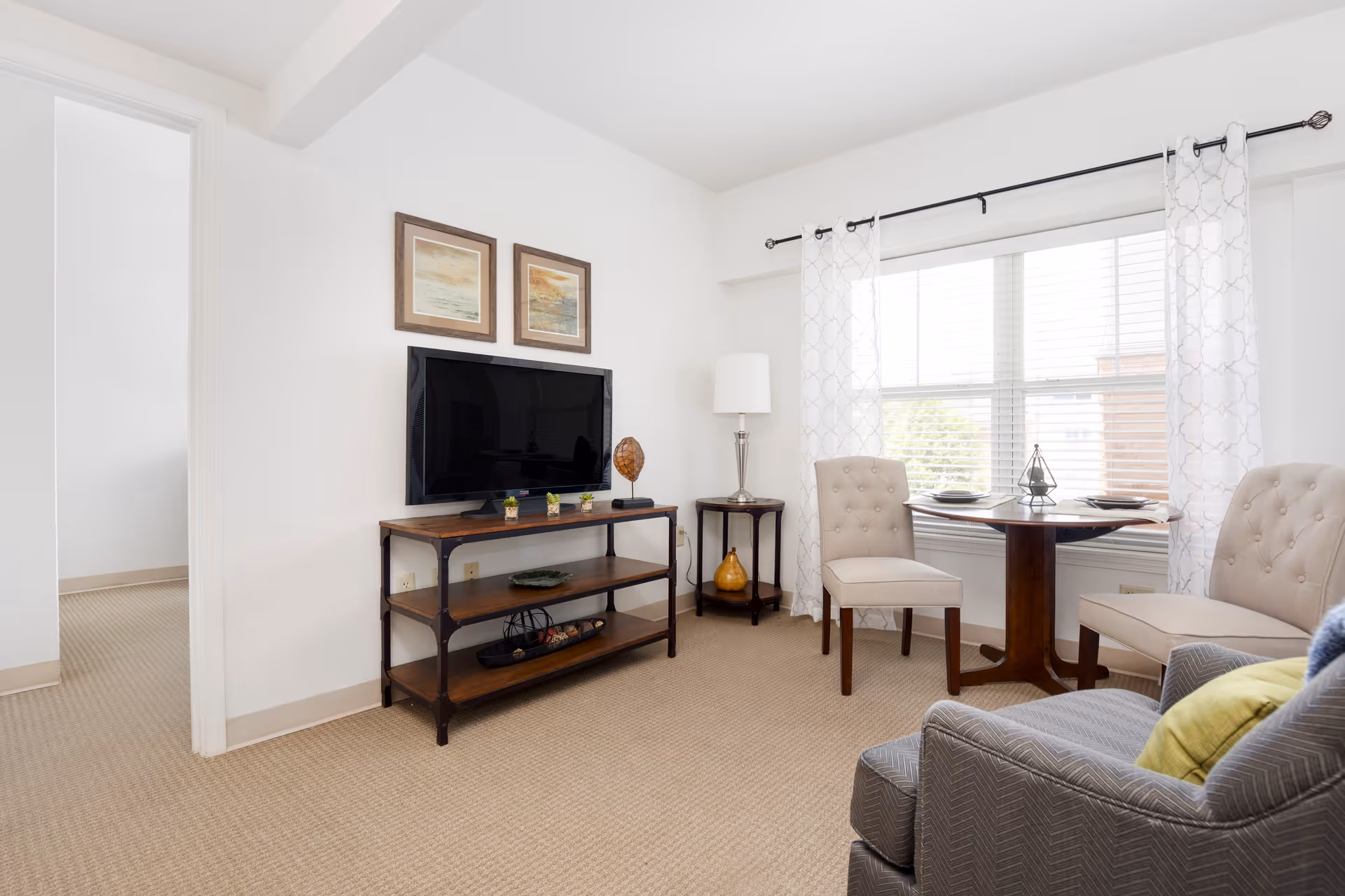 A bright living room with beige carpet and white walls featuring a flat-screen TV on a wooden stand, two framed pictures above the TV, a small round wooden table with two beige upholstered chairs near a window with white patterned curtains, and a gray armchair with a yellow pillow in the foreground.