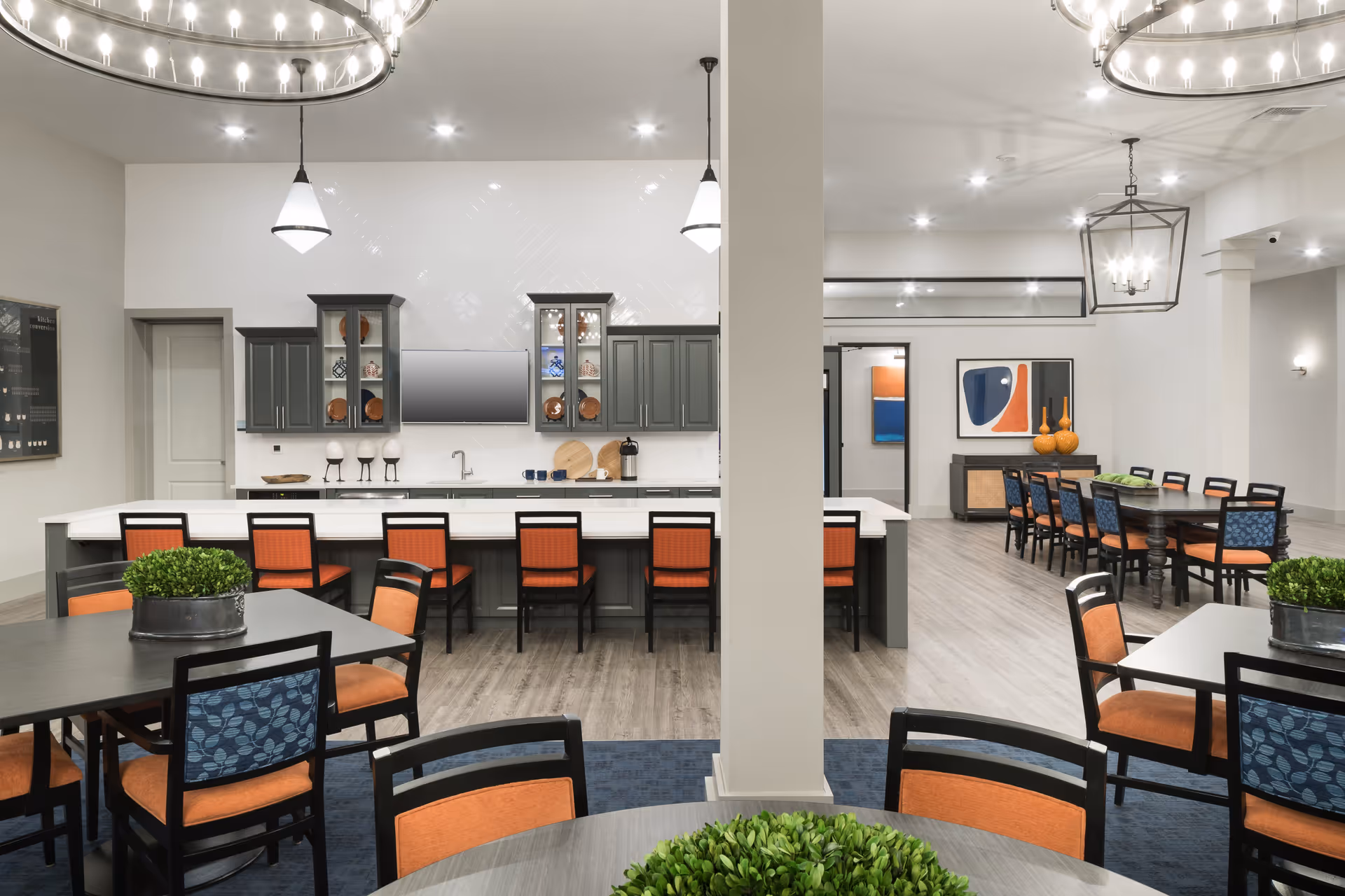 A modern communal dining and kitchen area in a senior living facility. The kitchen features gray cabinets, a white countertop island with orange cushioned chairs, and a wall-mounted TV. The dining area has multiple tables with black chairs that have orange and blue cushions. The room is well-lit with ceiling lights and decorative hanging fixtures, and there are green plants on the tables and abstract artwork on the walls.