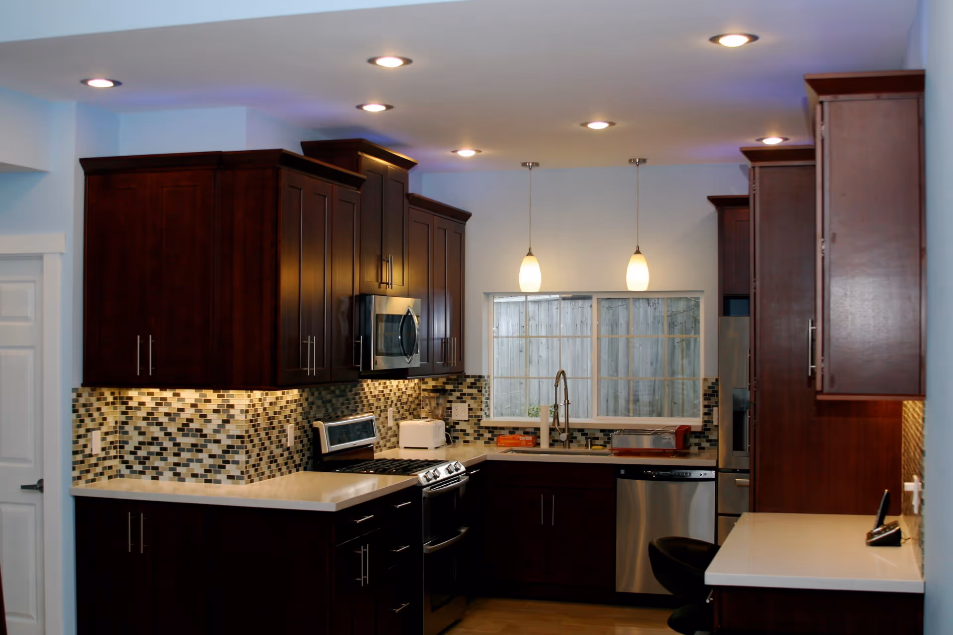 Modern kitchen with dark wooden cabinets, a mosaic tile backsplash, stainless steel appliances including a microwave, stove, and dishwasher. There is a window above the sink with two pendant lights hanging from the ceiling and recessed lighting illuminating the space.